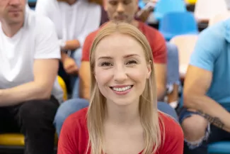 Blonde Frau in rotem Shirt, lächelt. Im Hintergrund sind weitere Personen zu sehen.