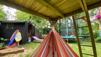 Blick aus einer bunten Hängematte auf einen sommerlichen Garten mit Trampolin, Gartenhaus und spielenden Kindern im Hintergrund. Blick aus einer bunten Hängematte auf einen sommerlichen Garten mit Trampolin, Gartenhaus und spielenden Kindern im Hintergrund.