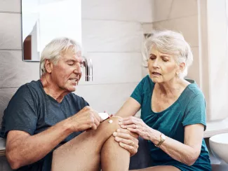 Älteres Ehepaar im Badezimmer: Der Mann cremt sein Knie mit Diclofenac-Gel ein, die Frau wischt das Gel mit einem Tuch ab und entsorgt es korrekt im Hausmüll – Motiv der Kampagne „Kein Diclofenac ins Abwasser!“.