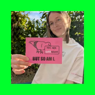Junge Studentin im Garten hält eine pinke Postkarte mit der Kreide-Taube und dem Spruch „You are gurrgeous — but so am I.“ in die Kamera.