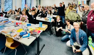 Ein voll besetzter Seminarraum: Über 60 Studierende und Lehrende posieren lachend und winkend für ein Gruppenfoto. Auf den Tischen liegen zahlreiche Design-Arbeitsmaterialien und Muster.