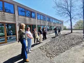 Mehrere Personen stehen vor dem Gebäude der ehemaligen Hochschule für Gestaltung Ulm; die Betonfassade mit langen Fensterbändern ist bei sonnigem Wetter zu sehen.