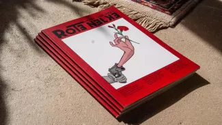 Vier Exemplare des Magazins Rote Nelken liegen gestapelt auf hellem Boden; auf dem roten Cover ragt aus einer Pistole eine Hand mit Nelke - ein Motiv für Widerstand, Fürsorge und politische Umkodierung.