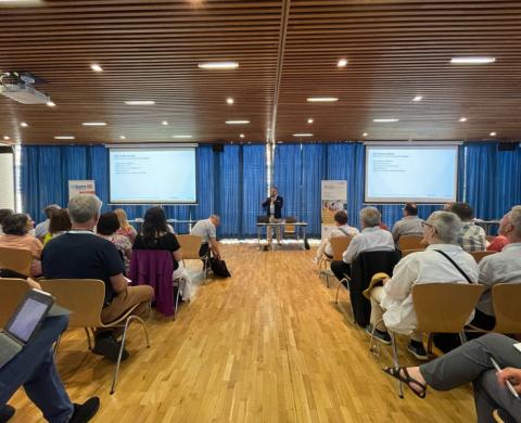 Konferenzraum mit Reihen aus Holzstühlen; vorne steht ein Redner am Pult zwischen zwei projizierten Folien, das Publikum hört zu.
