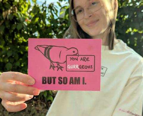 Junge Studentin im Garten hält eine pinke Postkarte mit der Kreide-Taube und dem Spruch „You are gurrgeous — but so am I.“ in die Kamera. Junge Studentin im Garten hält eine pinke Postkarte mit der Kreide-Taube und dem Spruch „You are gurrgeous — but so am I.“ in die Kamera.