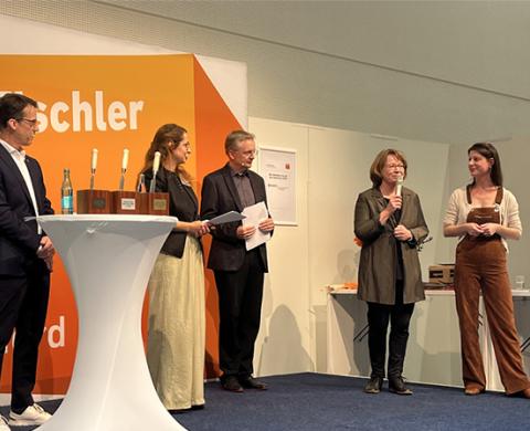 5 Menschen stehen auf einer Bühne und reden mit einem Mikro in der hand