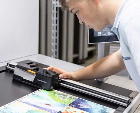 Ein Mann überprüft einen farbigen Druckbogen mit einem Messgerät auf einer Druckmaschine, um Farbgenauigkeit und Druckqualität zu kontrollieren.