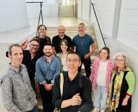 Eine Gruppe von Studierenden und Lehrenden steht lächelnd in einem hellen Treppenhaus des HfG-Gebäudes und blickt in die Kamera.