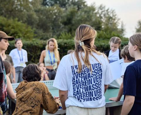 Eine Gruppe von Kindern und Jugendlichen steht im Freien um einen Tisch. Eine Person trägt ein weißes T-Shirt mit blauem SALZWASSER-VEREIN-Schriftzug auf dem Rücken.